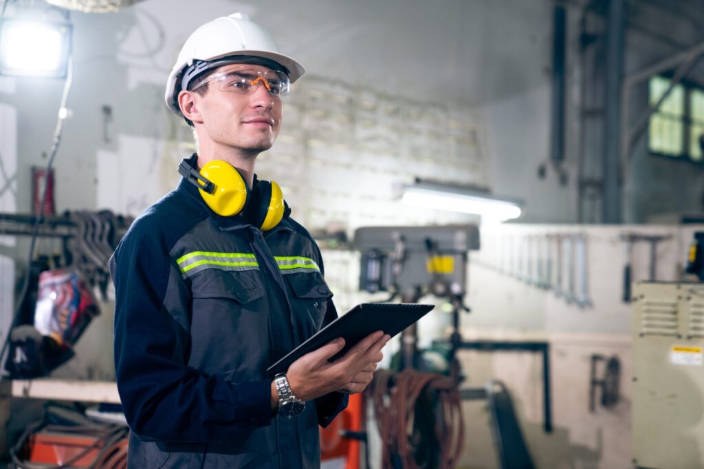 A safety-conscious worker in a hard hat and protective gear inspects a clipboard in a workshop, emphasizing safety and operational oversight.